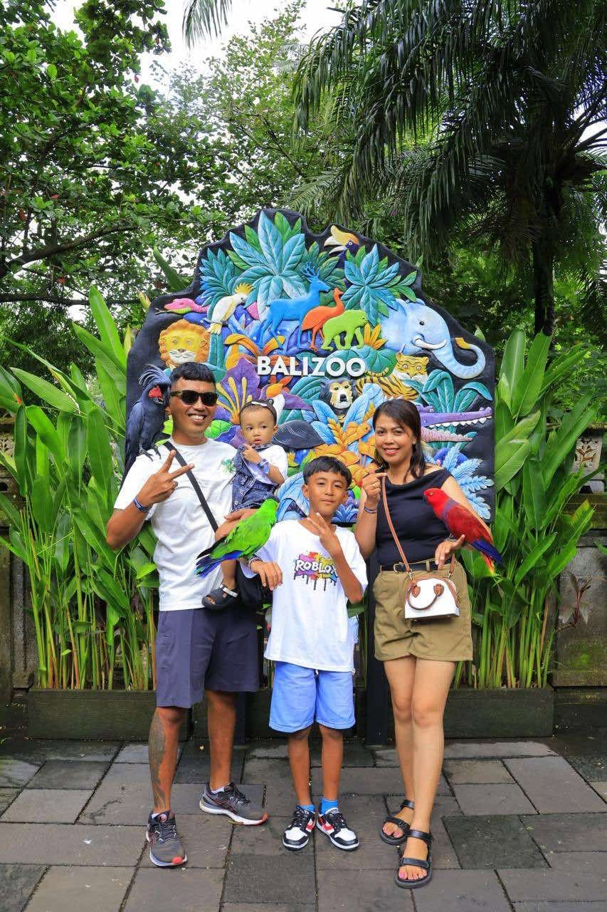 Anik and Made with their two children at the entrance to Bali Zoo, parrots perched on their arms.