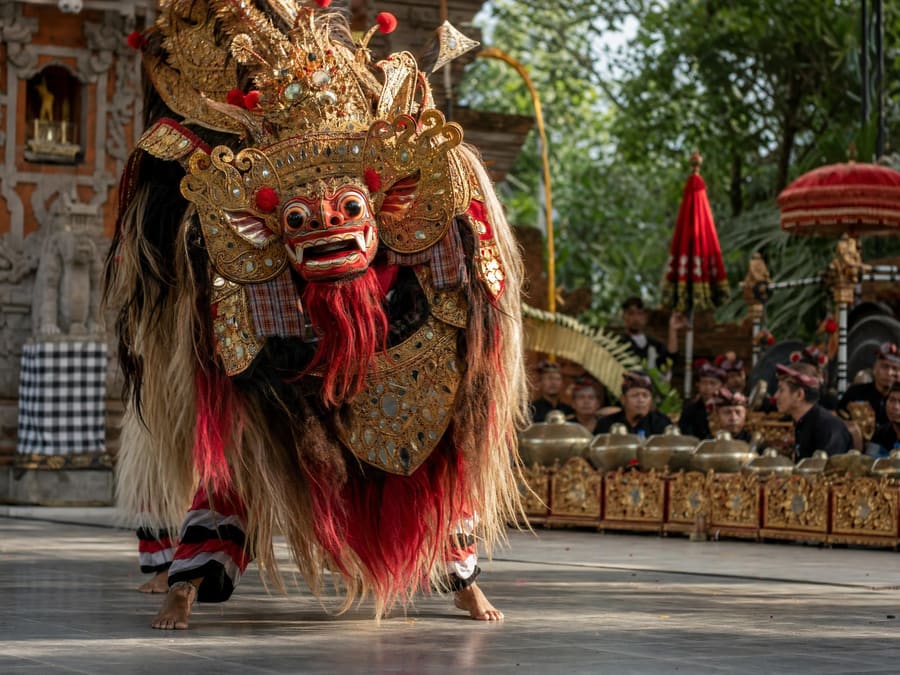 A Barong performer in an elaborate gilded lion mask with a red and gold beard, mid-performance on an open-air temple stage, a gamelan orchestra of bronze gongs blurred behind.