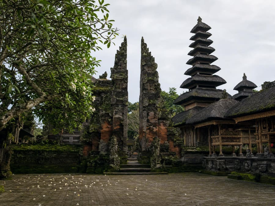 The quiet courtyard of an ancient Balinese village temple: a carved split gate, tiered black-thatch shrines and dark volcanic stone furred with green moss.