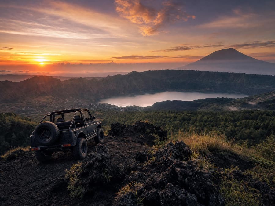 Sunrise over the Mount Batur caldera from a black lava ridge: an open-top 4WD jeep silhouetted in the foreground, the crater lake far below and Mount Agung on the horizon.