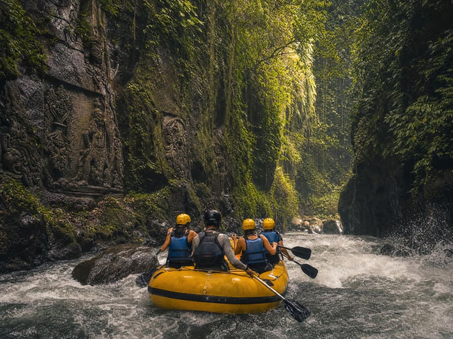 A yellow inflatable raft running gentle whitewater on the Ayung River in a deep jungle gorge, hand-carved stone relief panels visible in the canyon wall. The paddlers wear helmets and life jackets.