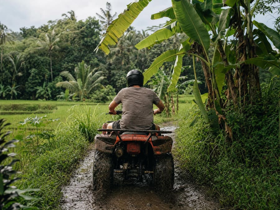 A quad bike on a muddy plantation track between rice fields and banana palms, mud splashed up the wheels. The rider wears a full helmet.