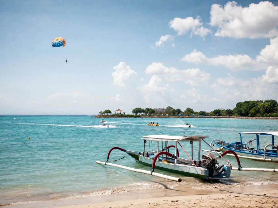 The calm, reef-sheltered turquoise bay at Tanjung Benoa: a parasail canopy high above the water, jet skis and a banana boat cutting white wakes far out, moored outrigger boats in the foreground.