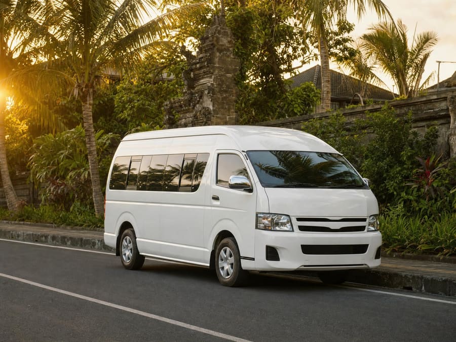 A clean white air-conditioned passenger minivan parked on a quiet palm-lined road at golden hour, dense tropical greenery and a stone temple wall behind.
