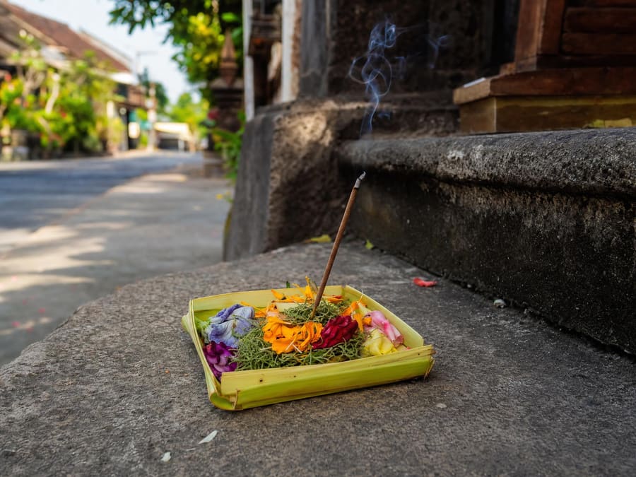 A canang sari offering — a small square tray woven from palm leaf, filled with coloured flower petals and a lit incense stick — resting on a pavement beside a shop step.