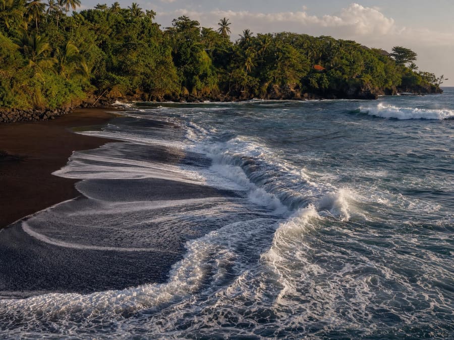 Strong surf breaking on a dark volcanic sand beach, with a rip current channel visible pulling seaward as a flatter, darker gap between the breaking waves.