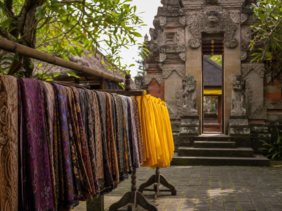 A rail of folded sarongs in traditional Balinese patterns with yellow sashes hanging beside them at a temple entrance, a carved stone gateway behind.