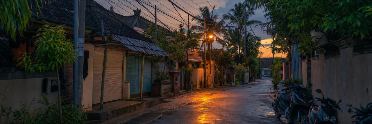 A narrow Bali side street at dusk — small shuttered shopfronts with awnings, tangled overhead cables, scooters parked along one side and a wet road reflecting the streetlight.