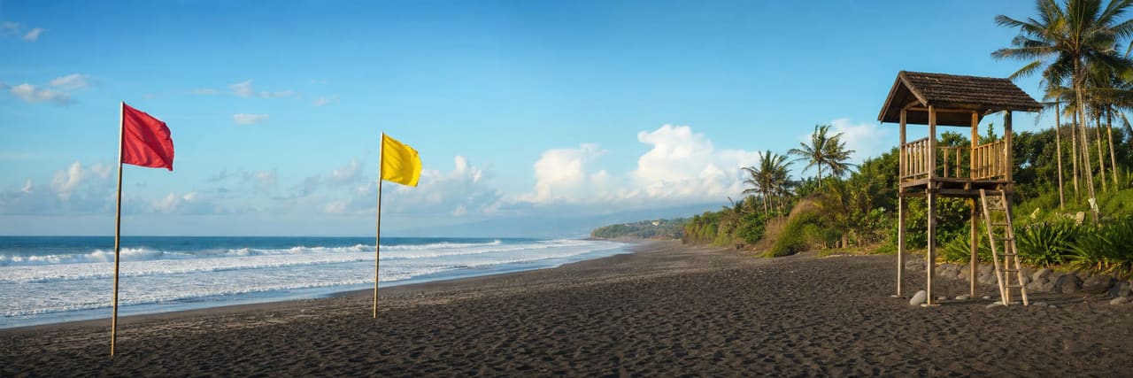 A patrolled Bali beach in the morning — a pair of red and yellow flags planted in dark volcanic sand marking the swimming zone, a wooden lifeguard tower to one side and breaking surf behind.